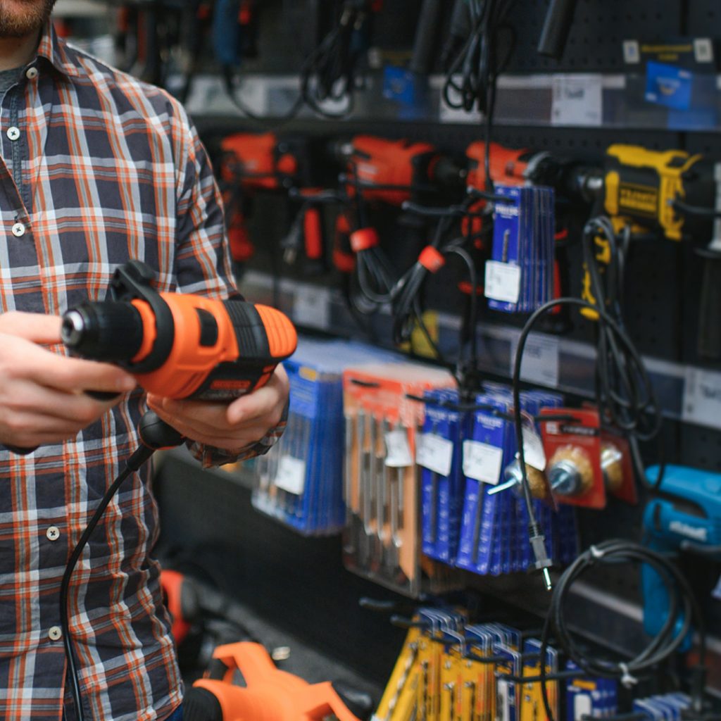 close-up of a man’s hands holding a drill at a hardware store shelf, inspecting the tool before purchase.